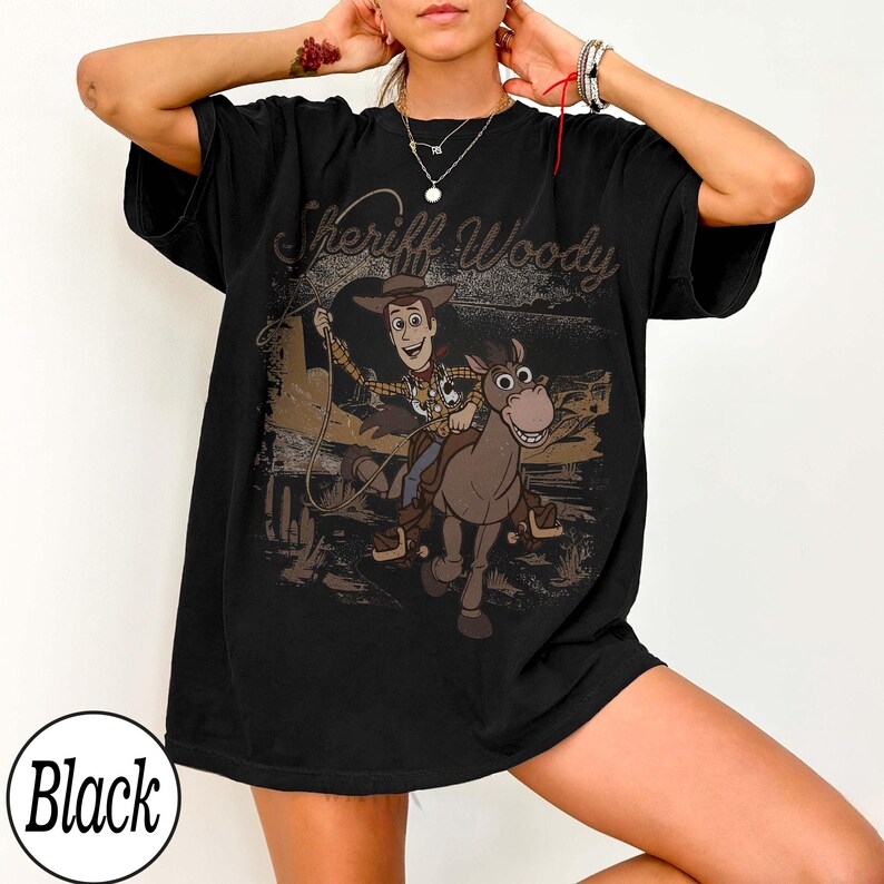 May include: Black t-shirt featuring a graphic of Sheriff Woody riding a horse, with the text "Sheriff Woody" in a brown, vintage font. The design is set against a desert landscape. The shirt is a relaxed fit.
