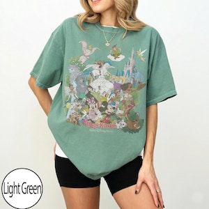 May include: Light green t-shirt featuring a colorful graphic of Disney characters, including Mickey Mouse, Dumbo, and Tinkerbell. The text "Magic Kingdom Walt Disney World" is printed at the bottom of the design. The shirt is a relaxed fit.
