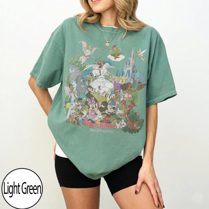 May include: Light green t-shirt featuring a colorful graphic of Disney characters, including Mickey Mouse, Dumbo, and Tinkerbell. The text "Magic Kingdom Walt Disney World" is printed at the bottom of the design. The shirt is a relaxed fit.