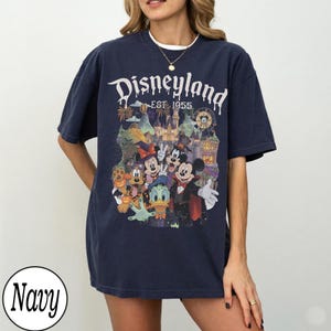 Puede incluir: Una camiseta azul marino de manga corta con un estampado gr&aacute;fico de personajes de Disney con disfraces de Halloween, incluyendo a Mickey Mouse, Minnie Mouse, Donald Duck y Pluto, con el texto Disneyland EST. 1955 y un castillo.