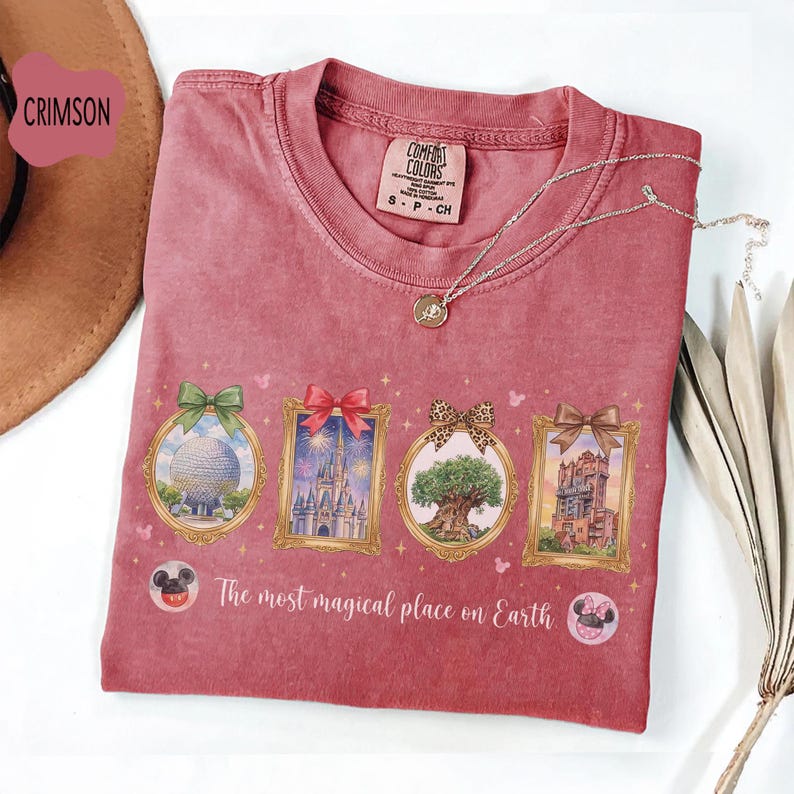 K&ouml;nnte beinhalten: Karmesinrotes Comfort Colors T-Shirt mit vier gerahmten Themenpark-Symbolen, darunter eine Kugel, ein Schloss, ein Baum und ein Turm, jeweils mit einer Schleife. Der Text "The most magical place on Earth" ist darunter gedruckt. Eine silberne Halskette liegt &uuml;ber dem T-Shirt.
