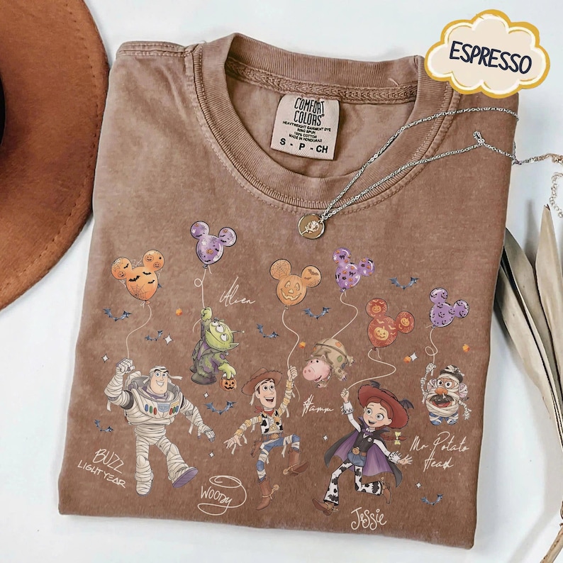 Pode incluir: Camiseta cor de caf&eacute; com personagens de Toy Story da Disney segurando bal&otilde;es em forma de Mickey Mouse. A camisa inclui Buzz Lightyear, Woody, Jessie e Sr. Cabe&ccedil;a de Batata. Um colar de prata com um pingente repousa sobre a camisa.