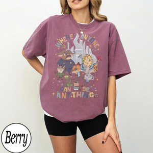 May include: Berry-coloured t-shirt with a colourful graphic featuring cartoon characters and the text "WHERE ANYONE CAN BE ANYTHING". The shirt has a crew neck and short sleeves.
