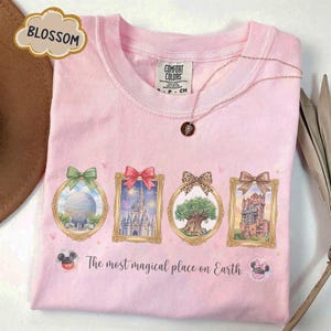 K&ouml;nnte beinhalten: Rosa Comfort Colors T-Shirt mit vier gerahmten Illustrationen von Themenpark-Wahrzeichen, jeweils mit einer Schleife versehen. Der Text "The most magical place on Earth" ist darunter aufgedruckt. Eine goldene Halskette mit Anh&auml;nger ist ebenfalls sichtbar.