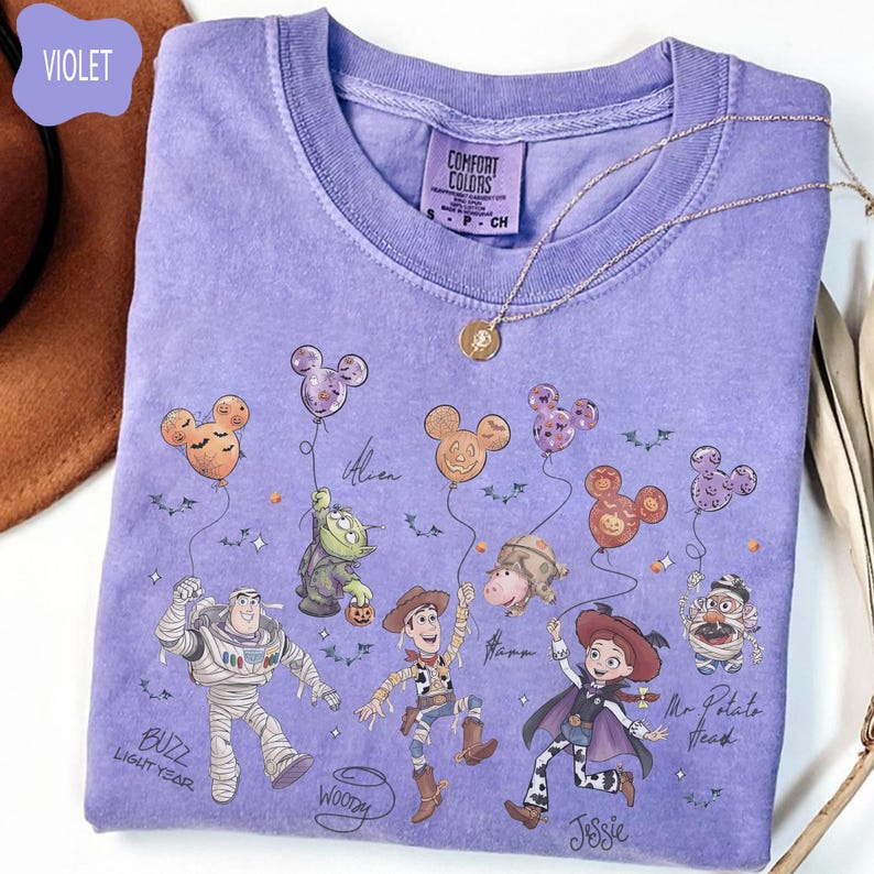 Pode incluir: Uma camiseta violeta Comfort Colors com personagens de Toy Story segurando bal&otilde;es do Mickey Mouse. Buzz Lightyear, Woody, Jessie e Sr. Cabe&ccedil;a de Batata s&atilde;o retratados. Um colar de ouro est&aacute; sobre a camiseta.