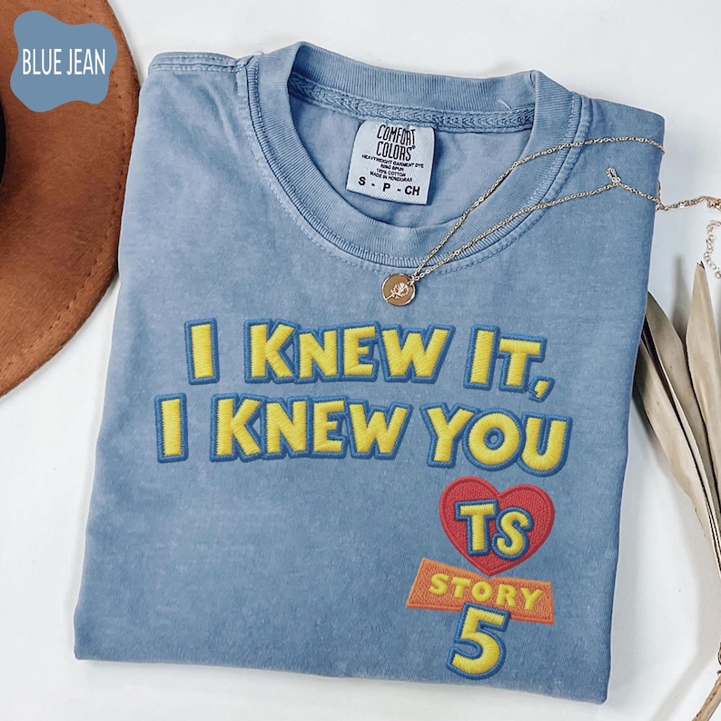 Puede incluir: Una camiseta color azul vaquero con el texto bordado en amarillo I KNEW IT, I KNEW YOU, un coraz&oacute;n rojo con las letras amarillas TS y el texto amarillo STORY 5. La camiseta tiene una etiqueta de la marca Comfort Colors y un collar dorado.