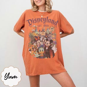 Puede incluir: Una camiseta naranja de manga corta con un estampado gr&aacute;fico de personajes de Disney con disfraces de Halloween frente a un castillo. El texto dice Disneyland Est. 1955. El dise&ntilde;o incluye a Mickey Mouse, Minnie Mouse, Donald Duck y Pluto.