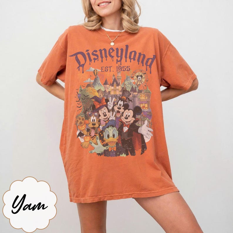 Puede incluir: Una camiseta naranja de manga corta con un estampado gr&aacute;fico de personajes de Disney con disfraces de Halloween frente a un castillo. El texto dice Disneyland Est. 1955. El dise&ntilde;o incluye a Mickey Mouse, Minnie Mouse, Donald Duck y Pluto.
