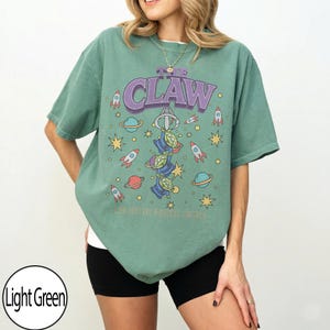 Puede incluir: Una camiseta de manga corta de color verde claro con un estampado gr&aacute;fico de tres alien&iacute;genas verdes colgados del gancho de una m&aacute;quina de garra. El texto THE CLAW aparece impreso encima, rodeado de cohetes, planetas y estrellas amarillas.