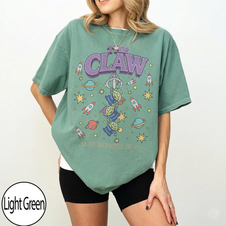 Puede incluir: Una camiseta de manga corta de color verde claro con un estampado gr&aacute;fico de tres alien&iacute;genas verdes colgados del gancho de una m&aacute;quina de garra. El texto THE CLAW aparece impreso encima, rodeado de cohetes, planetas y estrellas amarillas.