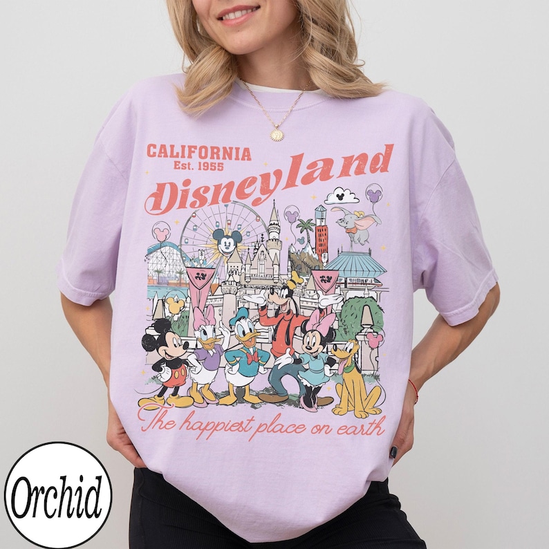 May include: A lilac t-shirt featuring a Disneyland graphic. The design includes Disney characters and park attractions, with text reading "California Disneyland Est. 1955" and "The happiest place on earth."