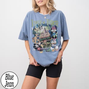 K&ouml;nnte beinhalten: Hellblaues T-Shirt mit einem Grafikdruck von Disney-Figuren und dem Text "Tower of Terror". Das Design zeigt Micky, Minnie, Donald und Goofy vor einem Spukhotel. Das T-Shirt hat eine hellblaue Farbe.