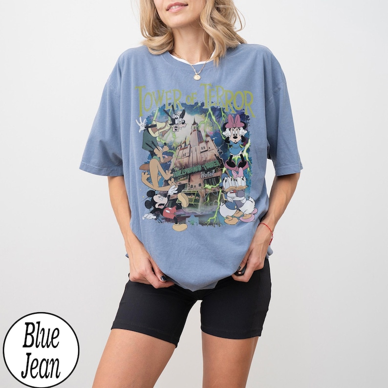 K&ouml;nnte beinhalten: Hellblaues T-Shirt mit einem Grafikdruck von Disney-Figuren und dem Text "Tower of Terror". Das Design zeigt Micky, Minnie, Donald und Goofy vor einem Spukhotel. Das T-Shirt hat eine hellblaue Farbe.