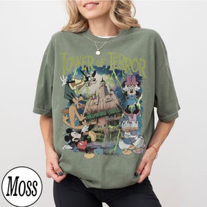 K&ouml;nnte beinhalten: Olivgr&uuml;nes T-Shirt mit einer Grafik von Disney-Figuren und dem Text "Tower of Terror". Das Design zeigt Micky Maus, Minnie Maus, Goofy, Donald Duck und Daisy Duck vor einem Spukhotel.