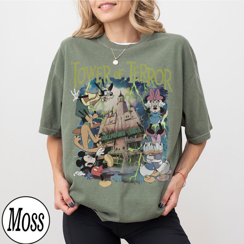 K&ouml;nnte beinhalten: Olivgr&uuml;nes T-Shirt mit einer Grafik von Disney-Figuren und dem Text "Tower of Terror". Das Design zeigt Micky Maus, Minnie Maus, Goofy, Donald Duck und Daisy Duck vor einem Spukhotel.