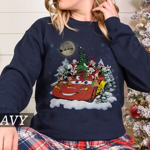 K&ouml;nnte beinhalten: Marineblaues Sweatshirt mit Weihnachtsgrafik. Das Design zeigt ein rotes Auto mit Disney-Figuren, einen Weihnachtsbaum und den Text "Merry Christmas". Schneeflocken und ein Vollmond mit dem Schlitten des Weihnachtsmanns erg&auml;nzen die festliche Szene.