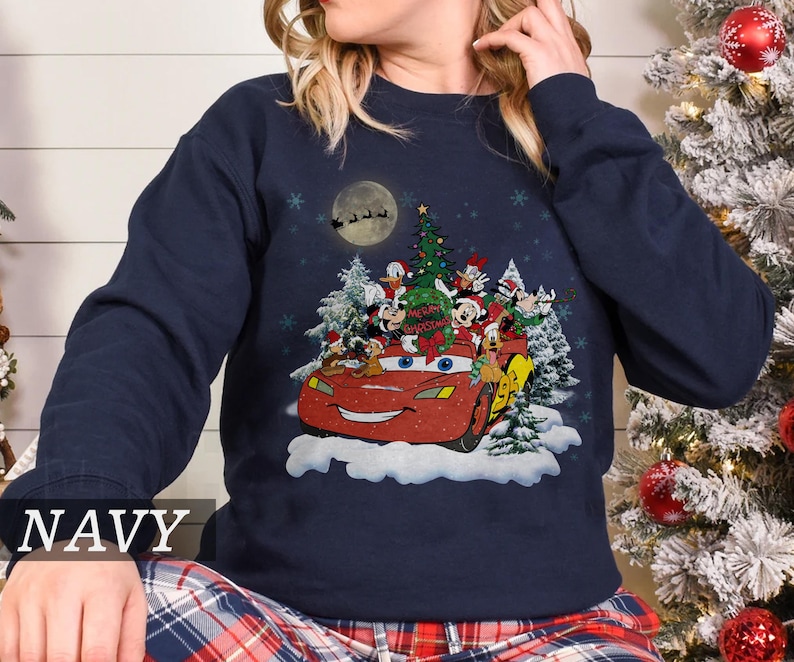 K&ouml;nnte beinhalten: Marineblaues Sweatshirt mit Weihnachtsgrafik. Das Design zeigt ein rotes Auto mit Disney-Figuren, einen Weihnachtsbaum und den Text "Merry Christmas". Schneeflocken und ein Vollmond mit dem Schlitten des Weihnachtsmanns erg&auml;nzen die festliche Szene.