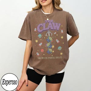 Puede incluir: Una camiseta marr&oacute;n de manga corta con un gr&aacute;fico de tres alien&iacute;genas verdes de tres ojos colgando de un gancho de m&aacute;quina de garra. El dise&ntilde;o incluye planetas, estrellas y cohetes con el texto THE CLAW y THE MYSTIC PORTAL AWAITS en morado y blanco.