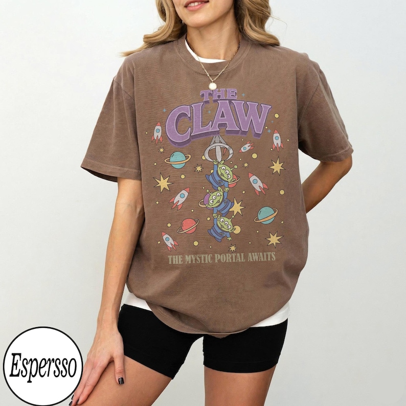 Puede incluir: Una camiseta marr&oacute;n de manga corta con un gr&aacute;fico de tres alien&iacute;genas verdes de tres ojos colgando de un gancho de m&aacute;quina de garra. El dise&ntilde;o incluye planetas, estrellas y cohetes con el texto THE CLAW y THE MYSTIC PORTAL AWAITS en morado y blanco.