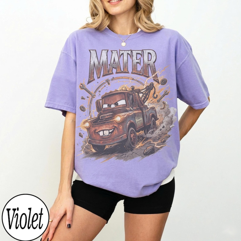 May include: Violet-colored t-shirt featuring a graphic of Mater from the movie Cars. The design includes the word "MATER" in a distressed font above the character. The shirt has a relaxed fit and short sleeves.