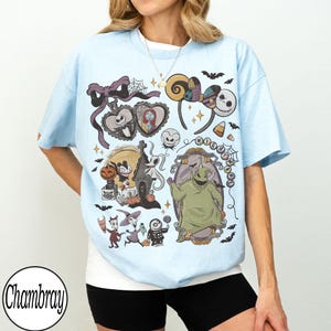 May include: Light blue t-shirt featuring a colorful graphic design with characters and elements from "The Nightmare Before Christmas." The design includes Jack Skellington, Sally, Oogie Boogie, and other iconic imagery. The shirt has a crew neck and short sleeves.