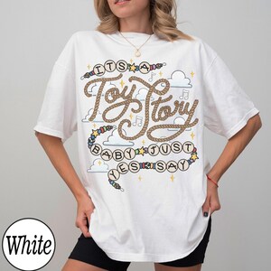 May include: A white short-sleeved t-shirt featuring a graphic design with the text "ITS A Toy Story BABY JUST YES SAY". The design includes brown rope-style lettering, colorful beads, small yellow stars, musical notes, and blue cloud illustrations.
