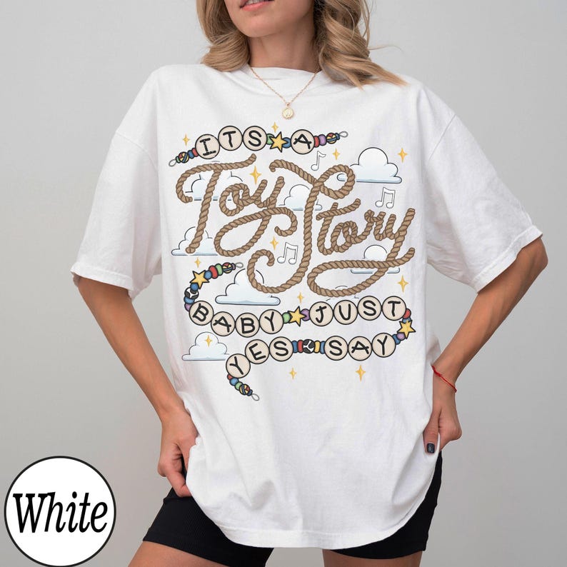 May include: A white short-sleeved t-shirt featuring a graphic design with the text "ITS A Toy Story BABY JUST YES SAY". The design includes brown rope-style lettering, colorful beads, small yellow stars, musical notes, and blue cloud illustrations.