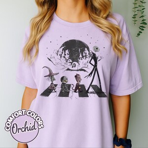 Peut inclure: T-shirt Comfort Colors lilas avec un motif de personnages traversant une rue, sur fond de pleine lune et de ville effrayante. Le t-shirt porte les mots "Comfort Colors Orchid" dans un cercle en bas &agrave; gauche.