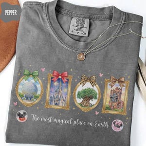 K&ouml;nnte beinhalten: Ein graues Comfort Colors T-Shirt mit vier gerahmten Illustrationen, die jeweils mit einer Schleife verziert sind und einen Globus, ein Schloss, einen Baum und ein Geb&auml;ude darstellen. Der Text "The most magical place on Earth" ist darunter aufgedruckt. Eine silberne Halskette liegt auf dem T-Shirt.