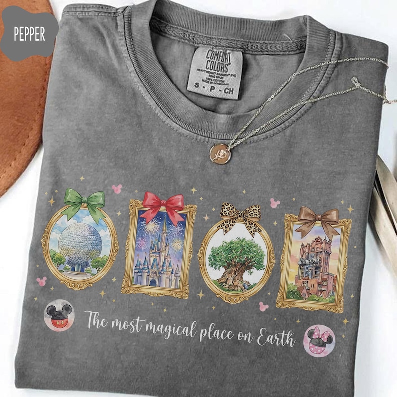 K&ouml;nnte beinhalten: Ein graues Comfort Colors T-Shirt mit vier gerahmten Illustrationen, die jeweils mit einer Schleife verziert sind und einen Globus, ein Schloss, einen Baum und ein Geb&auml;ude darstellen. Der Text "The most magical place on Earth" ist darunter aufgedruckt. Eine silberne Halskette liegt auf dem T-Shirt.