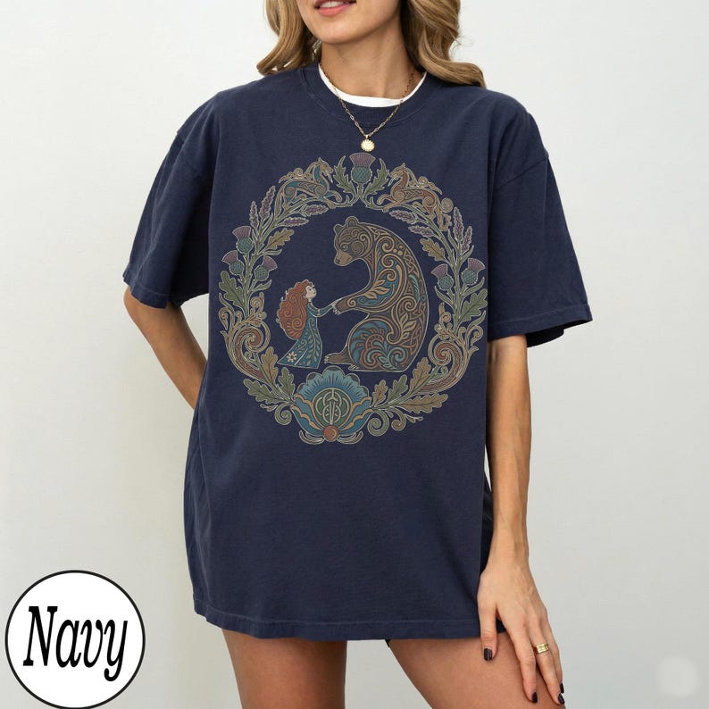 以下が含まれることがあります： ネイビーブルーのTシャツ。円形のデザインで、クマと女の子が花のリースに囲まれています。デザインには茶色、緑、青の色合いが使われています。「Navy」の文字が左下に白い円で印刷されています。
