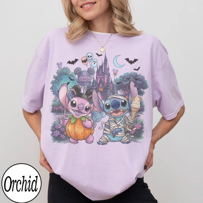 May include: A light purple t-shirt featuring a graphic print of two blue and pink alien characters in Halloween costumes. One character is dressed as a pumpkin, the other as a mummy holding an ice cream cone. The background shows a castle, bats, and balloons.