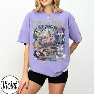 K&ouml;nnte beinhalten: Ein violettes T-Shirt mit einer Grafik von Disney-Figuren und dem Text "Tower of Terror". Das Design umfasst Micky Maus, Minnie Maus, Goofy und Donald Duck. Das Shirt hat eine lockere Passform.