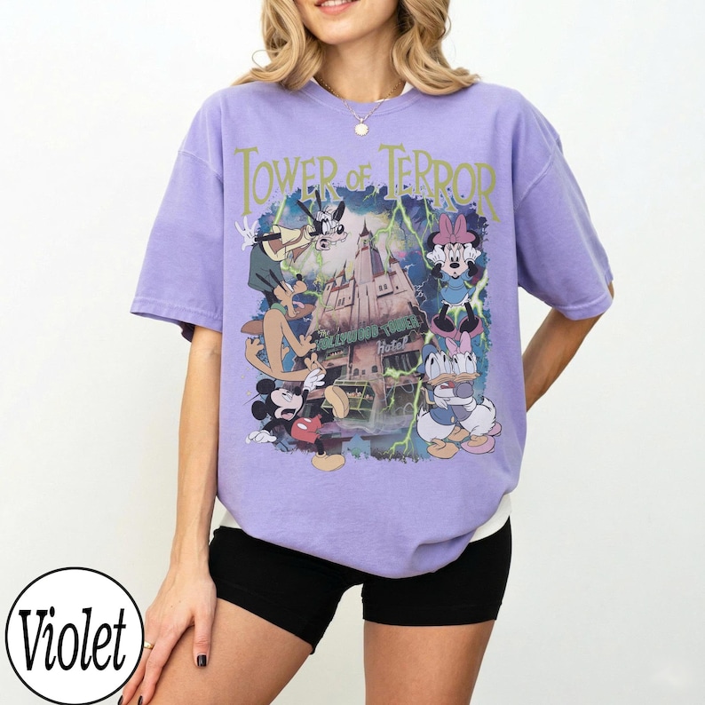 K&ouml;nnte beinhalten: Ein violettes T-Shirt mit einer Grafik von Disney-Figuren und dem Text "Tower of Terror". Das Design umfasst Micky Maus, Minnie Maus, Goofy und Donald Duck. Das Shirt hat eine lockere Passform.