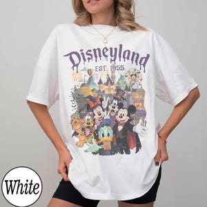 Puede incluir: Una camiseta blanca con un estampado gr&aacute;fico de Disneyland y el texto "Disneyland EST. 1955". El dise&ntilde;o incluye ilustraciones de Mickey Mouse, Minnie Mouse, Donald Duck y Pluto con disfraces de Halloween frente a un castillo.