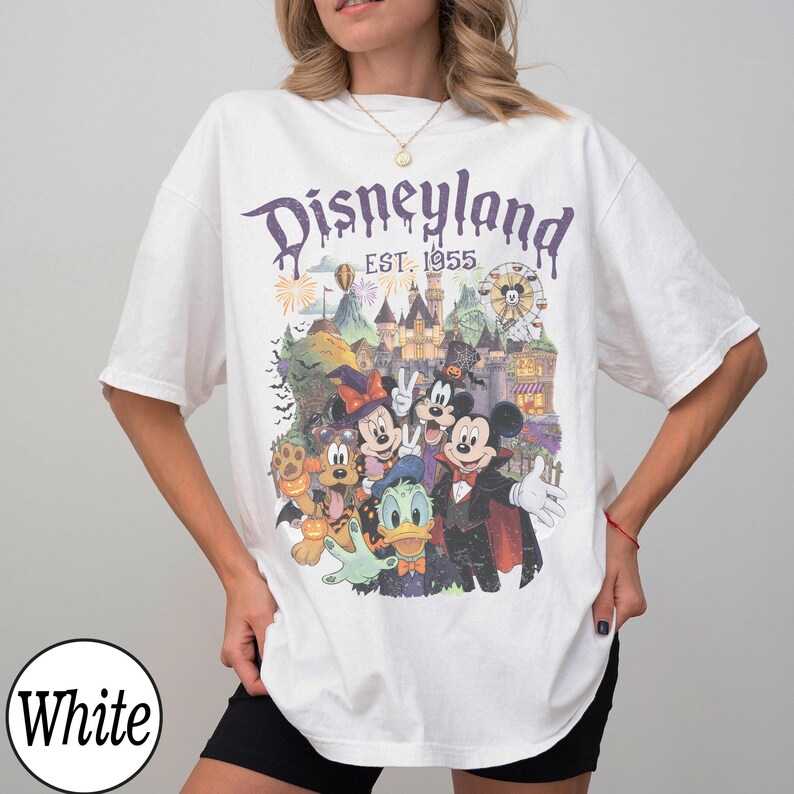 Puede incluir: Una camiseta blanca con un estampado gr&aacute;fico de Disneyland y el texto "Disneyland EST. 1955". El dise&ntilde;o incluye ilustraciones de Mickey Mouse, Minnie Mouse, Donald Duck y Pluto con disfraces de Halloween frente a un castillo.