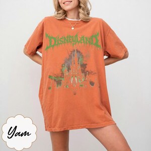 May include: Orange t-shirt with the word "Disneyland" in green, stylized text above a castle graphic. The shirt is a short-sleeved, oversized fit. The shirt is a solid orange color.