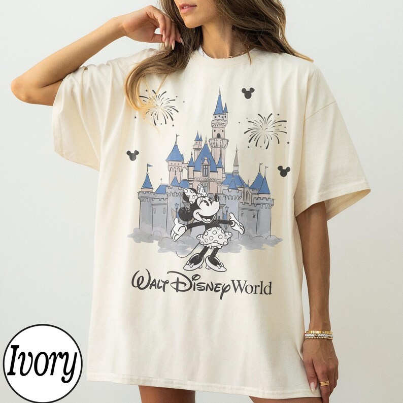 May include: Ivory-coloured t-shirt with a graphic of Minnie Mouse in front of a castle, fireworks, and the text "Walt Disney World." The shirt has short sleeves and a relaxed fit, suitable for casual wear.