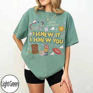 May include: A light green short-sleeve t-shirt featuring a graphic print with the text "I KNEW IT I KNEW YOU" and "infinity and beyond". The design includes illustrations of cowboy boots, alphabet blocks, a red hat, a star, and a beaded bracelet.