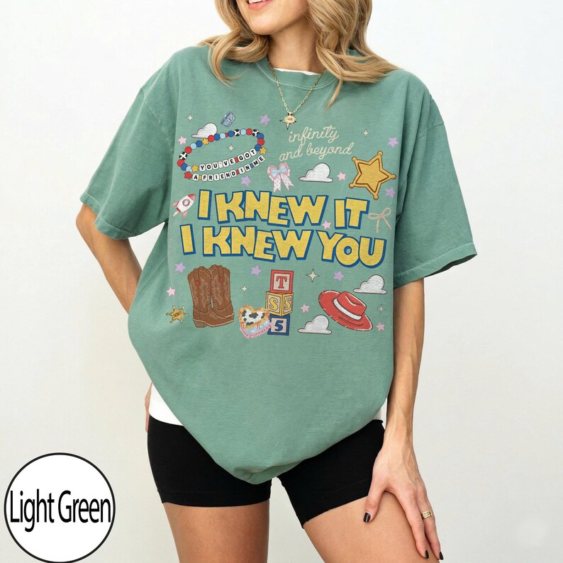 May include: A light green short-sleeve t-shirt featuring a graphic print with the text "I KNEW IT I KNEW YOU" and "infinity and beyond". The design includes illustrations of cowboy boots, alphabet blocks, a red hat, a star, and a beaded bracelet.