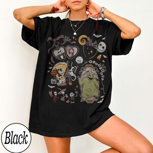 May include: Black t-shirt featuring a colorful graphic design with characters and elements from "The Nightmare Before Christmas." The design includes Jack Skellington, Sally, Oogie Boogie, and other iconic imagery. The word "Black" is printed on the bottom left.