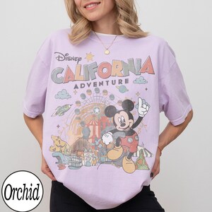 May include: A lilac-colored t-shirt featuring a graphic of Mickey Mouse with the text "Disney California Adventure". The design includes a Ferris wheel, rocket ship, and other amusement park elements. The shirt has short sleeves and a crew neck.