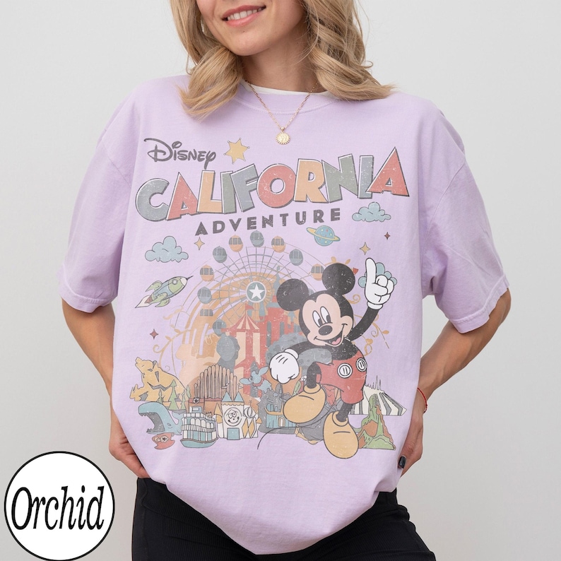 May include: A lilac-colored t-shirt featuring a graphic of Mickey Mouse with the text "Disney California Adventure". The design includes a Ferris wheel, rocket ship, and other amusement park elements. The shirt has short sleeves and a crew neck.