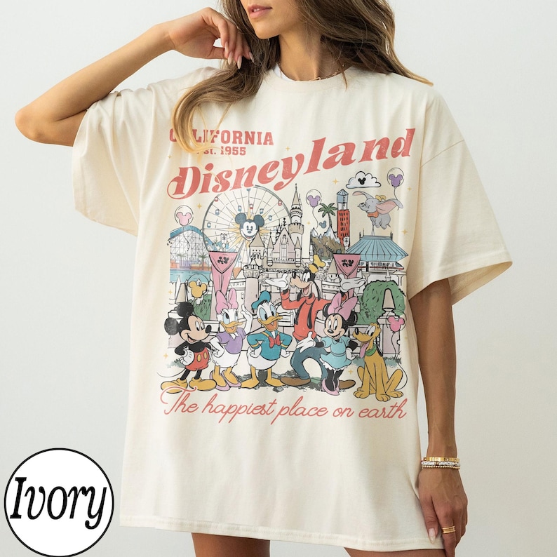 May include: Ivory-coloured t-shirt with a Disneyland graphic. The design showcases the Disneyland logo, park imagery, and Disney characters. Text includes "California Disneyland est. 1955" and "The happiest place on earth."