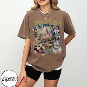 K&ouml;nnte beinhalten: Braunes T-Shirt mit einer Grafik von Disney-Figuren, darunter Micky Maus, Goofy und Minnie Maus, mit dem Text "Tower of Terror" und "Hollywood Tower Hotel". Das Shirt hat einen Vintage-Look.