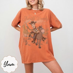 May include: Orange t-shirt featuring Sheriff Woody riding a horse, with the text "Sheriff Woody" above the graphic. The shirt has short sleeves and a relaxed fit. The word "Yam" is at the bottom left.