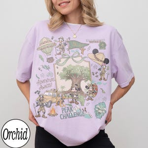 May include: A lilac t-shirt with a vibrant graphic of cartoon characters in safari gear. The design includes a hot air balloon, a jeep, a campfire, and the text "Adventure Awaits!" and "Peak Challenge."