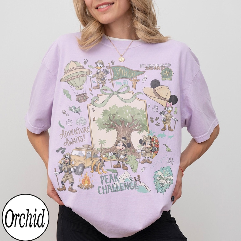 May include: A lilac t-shirt with a vibrant graphic of cartoon characters in safari gear. The design includes a hot air balloon, a jeep, a campfire, and the text "Adventure Awaits!" and "Peak Challenge."