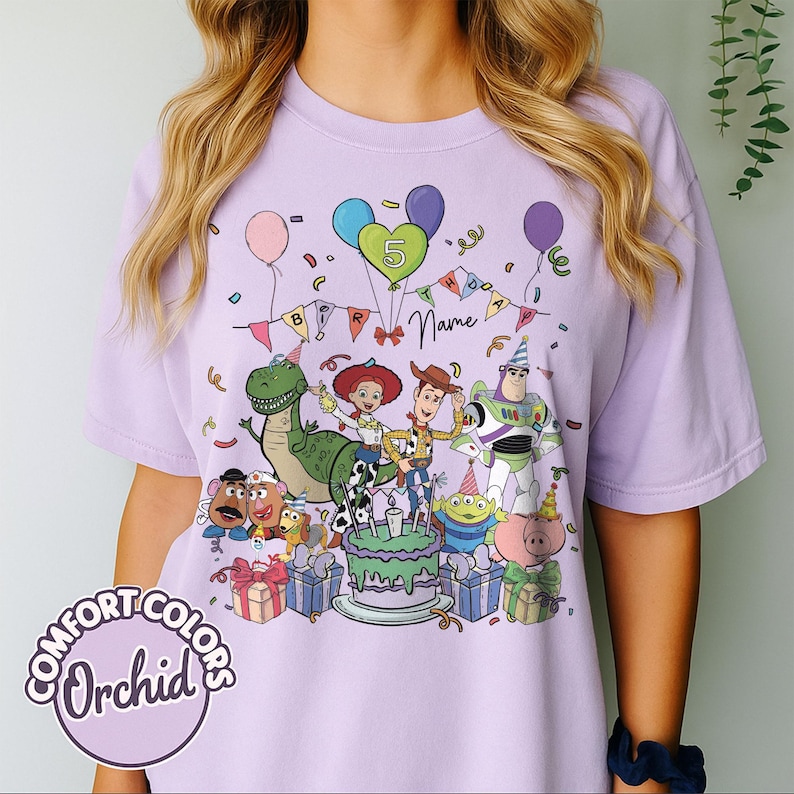 May include: A lilac Comfort Colors t-shirt featuring a colorful graphic of Toy Story characters celebrating a birthday. The design includes a cake, balloons, confetti, and the number 5. The word "Name" is also visible.