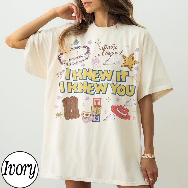 May include: An ivory t-shirt featuring a graphic print with the text "I KNEW IT I KNEW YOU" and "infinity and beyond". The design includes illustrations of cowboy boots, a red hat, alphabet blocks, a star, clouds, and a beaded necklace.